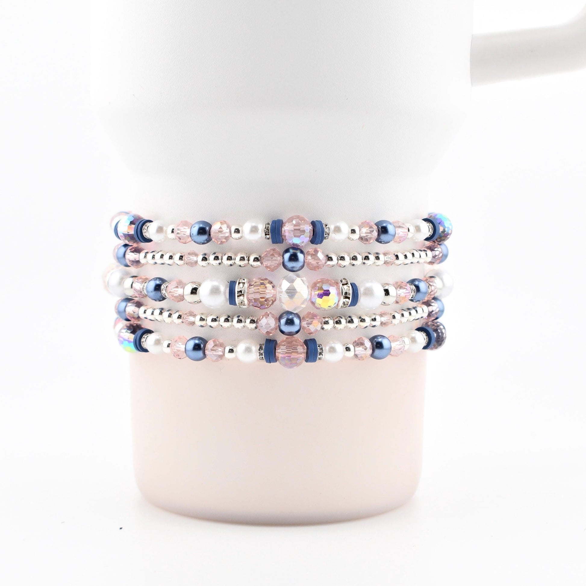 A white tumbler with several beaded bracelets wrapped around it. The bracelets feature a mix of pink, blue, and white beads, with silver accents. The beads are arranged in a repeating pattern, creating a decorative accessory.