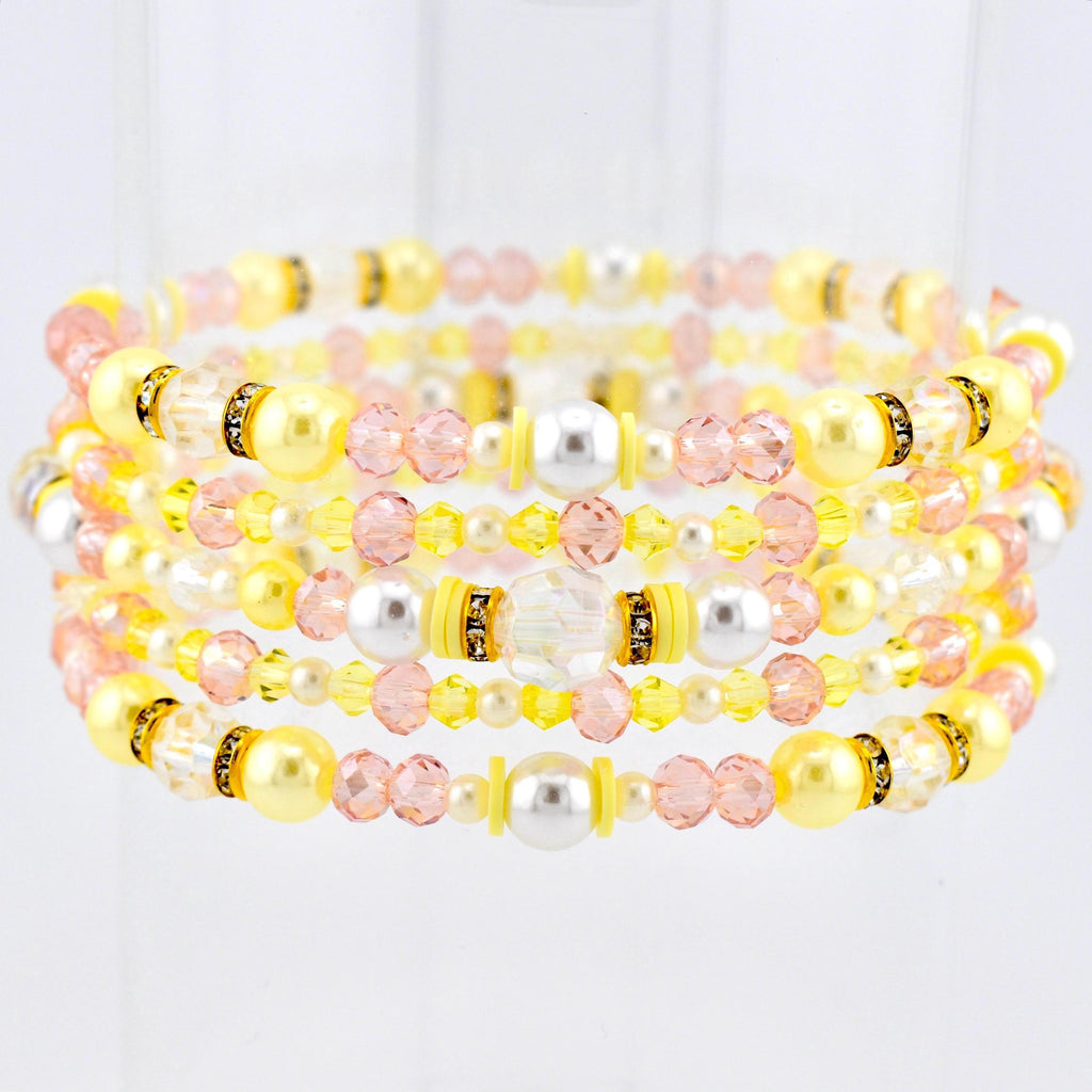 Stack of beaded bracelets in shades of yellow, pink, and white. The bracelets feature a variety of beads, including pearls, crystals, and smaller accent beads. Some sections have gold-colored spacers. The bracelets are stacked together.