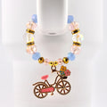 A beaded bracelet with a bicycle charm. The bracelet features clear, pink, and blue beads, with gold accents. The bicycle charm is pink with a basket of flowers and gold detailing.