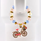 A beaded bracelet with a bicycle charm. The bracelet features clear, pink, and blue beads, with gold accents. The bicycle charm is pink with a basket of flowers and gold detailing.