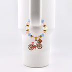A beaded drink charm with a bicycle pendant. The charm features light blue and pink beads, gold accents, and a bicycle pendant with a pink seat and a basket of flowers. The charm is attached to a white tumbler.