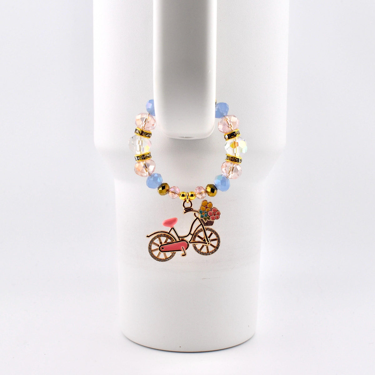 A beaded drink charm with a bicycle pendant. The charm features light blue and pink beads, gold accents, and a bicycle pendant with a pink seat and a basket of flowers. The charm is attached to a white tumbler.