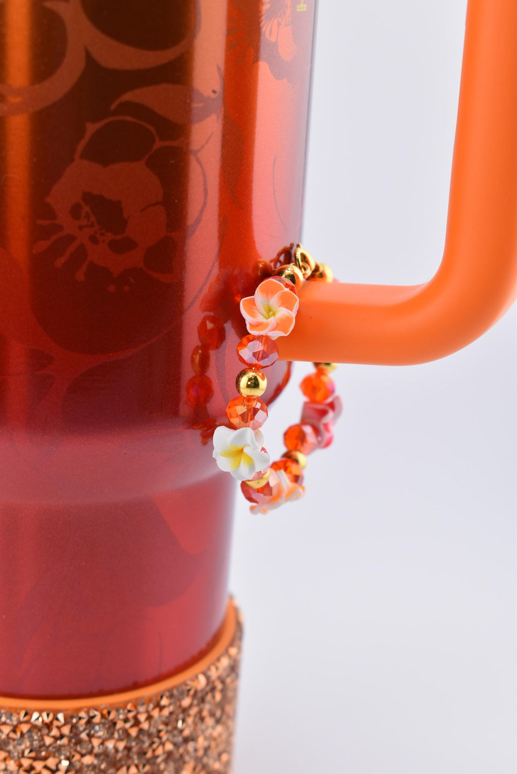 A Karol G Stanley with an orange handle and a decorative bracelet. The bracelet is adorned with orange, red, and gold beads, and small white and orange flower accents. The base of the tumbler has a copper-colored, glittery band.