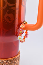 A Karol G Stanley with an orange handle and a decorative bracelet. The bracelet is adorned with orange, red, and gold beads, and small white and orange flower accents. The base of the tumbler has a copper-colored, glittery band.