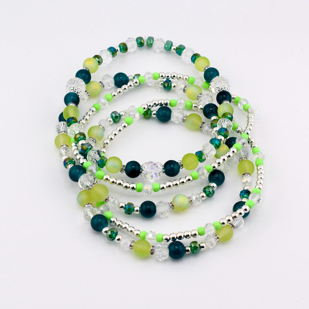 A collection of beaded bracelets in shades of green, teal, and clear. The bracelets feature a mix of round and faceted beads, interspersed with silver-colored spacers. The bracelets are arranged in a layered, circular pattern.