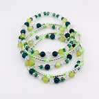 A collection of beaded bracelets in shades of green, teal, and clear. The bracelets feature a mix of round and faceted beads, interspersed with silver-colored spacers. The bracelets are arranged in a layered, circular pattern.