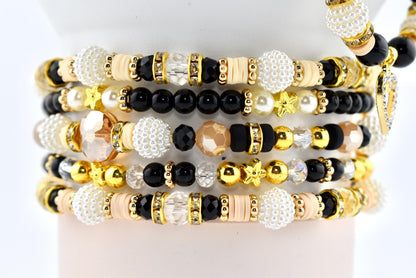 Stack of beaded bracelets in various designs. The bracelets feature a mix of black, white, gold, and clear beads. Some have star-shaped accents, while others include crystal and pearl details. The bracelets are stacked on a white surface.