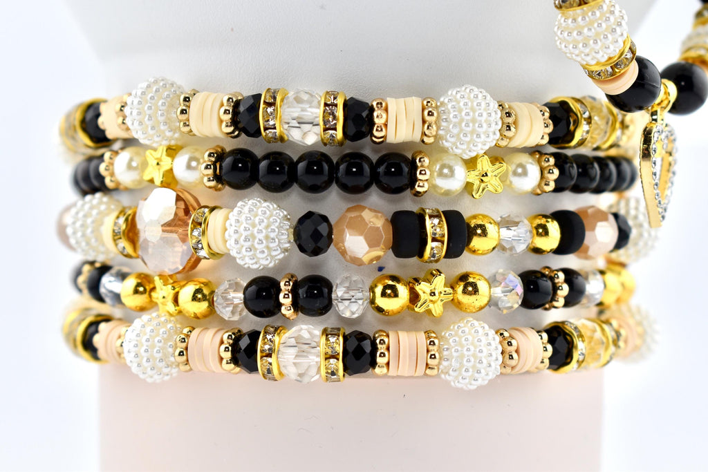 Stack of beaded bracelets in various designs. The bracelets feature a mix of black, white, gold, and clear beads. Some have star-shaped accents, while others include crystal and pearl details. The bracelets are stacked on a white surface.