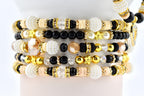 Stack of beaded bracelets in various designs. The bracelets feature a mix of black, white, gold, and clear beads. Some have star-shaped accents, while others include crystal and pearl details. The bracelets are stacked on a white surface.
