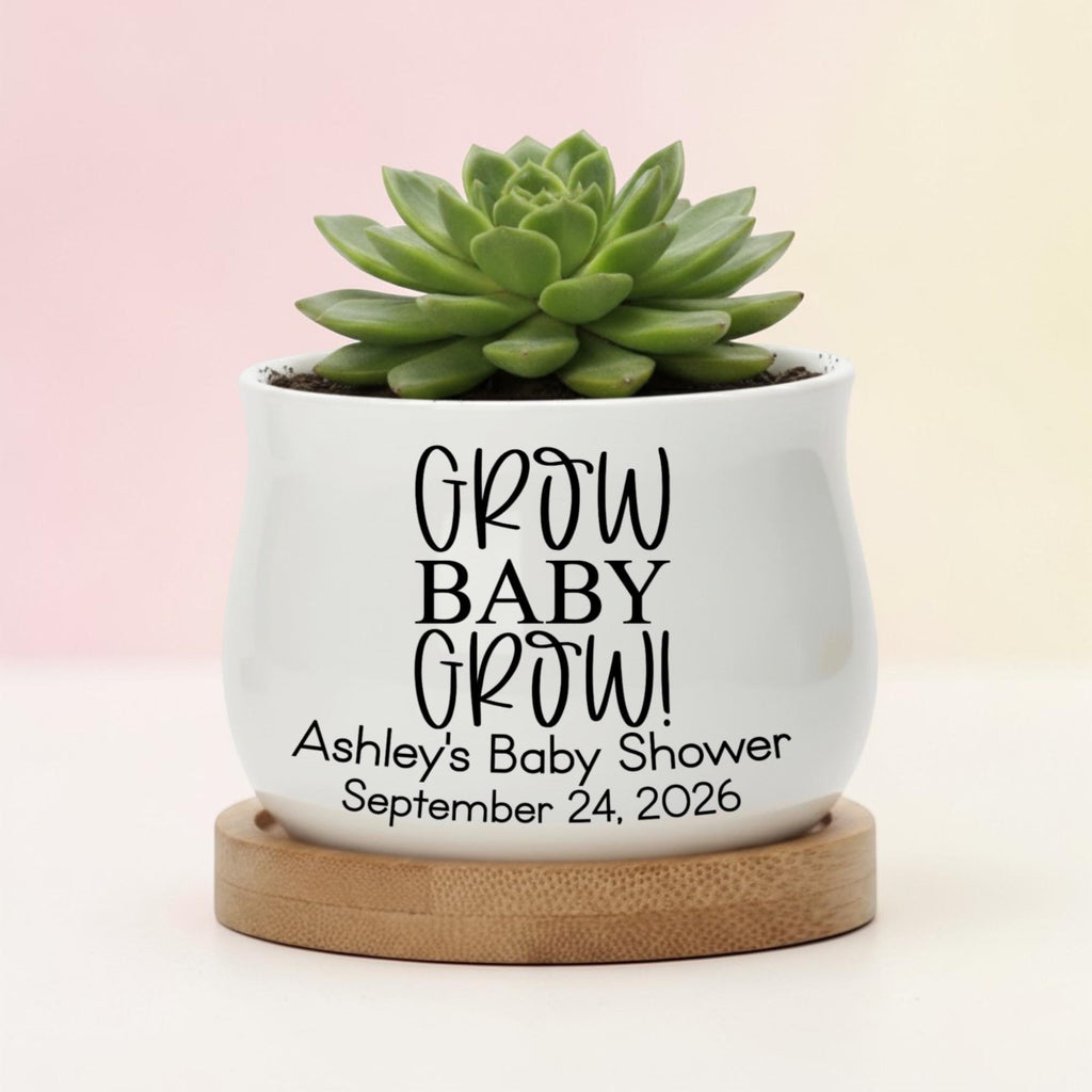 A white ceramic pot with the text "GROW BABY GROW!" and "Ashley's Baby Shower September 24, 2026" printed in black. The pot holds a green succulent plant and sits on a round wooden base.