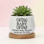 A white ceramic pot with the text "GROW BABY GROW!" and "Ashley's Baby Shower September 24, 2026" printed in black. The pot holds a green succulent plant and sits on a round wooden base.