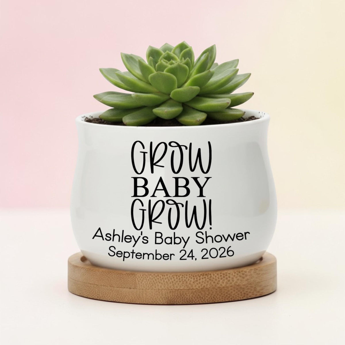 A white ceramic pot with the text "GROW BABY GROW!" and "Ashley's Baby Shower September 24, 2026" printed in black. The pot holds a green succulent plant and sits on a round wooden base.