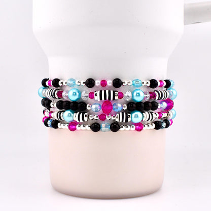Stack of beaded bracelets in shades of black, pink, blue, and silver. The bracelets feature a variety of beads, including small black and pink beads, larger blue beads, and silver accents. Some bracelets have striped beads.