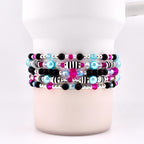 Stack of beaded bracelets in shades of black, pink, blue, and silver. The bracelets feature a variety of beads, including small black and pink beads, larger blue beads, and silver accents. Some bracelets have striped beads.