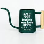 Emerald green metal gooseneck watering can with a wood handle. The watering can has a white vinyl design on the side that features a floral design and the text 'letting our love grow' and on the next two lines 'keith and kathy est. 2026'.