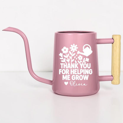A pink watering can with a long spout and wooden handle. The can features white floral and watering can graphics, along with the text "THANK YOU FOR HELPING ME GROW" and a name. Ideal for gardening.