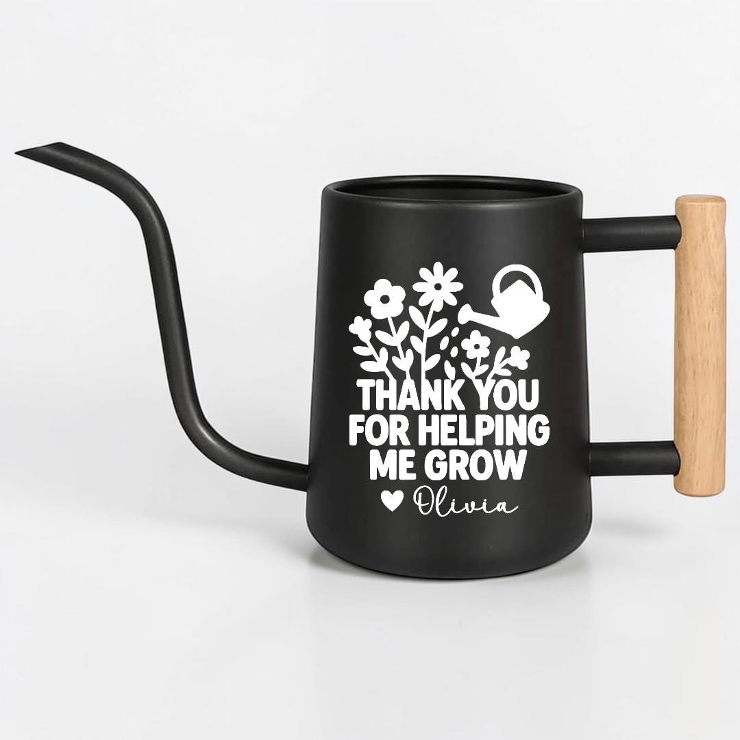 Black watering can with a long, curved spout and a wooden handle. The can features a white floral design and the text "THANK YOU FOR HELPING ME GROW" with the name "Olivia".