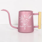 A dusty pink metal watering can with a long, slender spout and a light-colored wooden handle. The front of the watering can features a white line-art illustration of a floral bouquet above the printed words Custom Text.