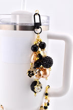 A decorative charm with a black clasp, gold chain, and various beads. The charm features black, gold, and clear beads, including a black rose, a faceted crystal, and a textured black ball. The charm is attached to a white mug.