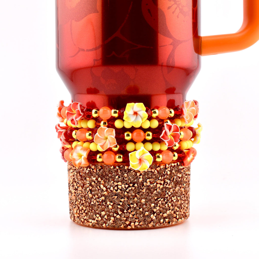 A Karol G Stanley with an orange handle, adorned with a floral beaded design. The design includes yellow, orange, and red beads, along with small flower accents. The base of the mug is covered in copper-colored glitter.