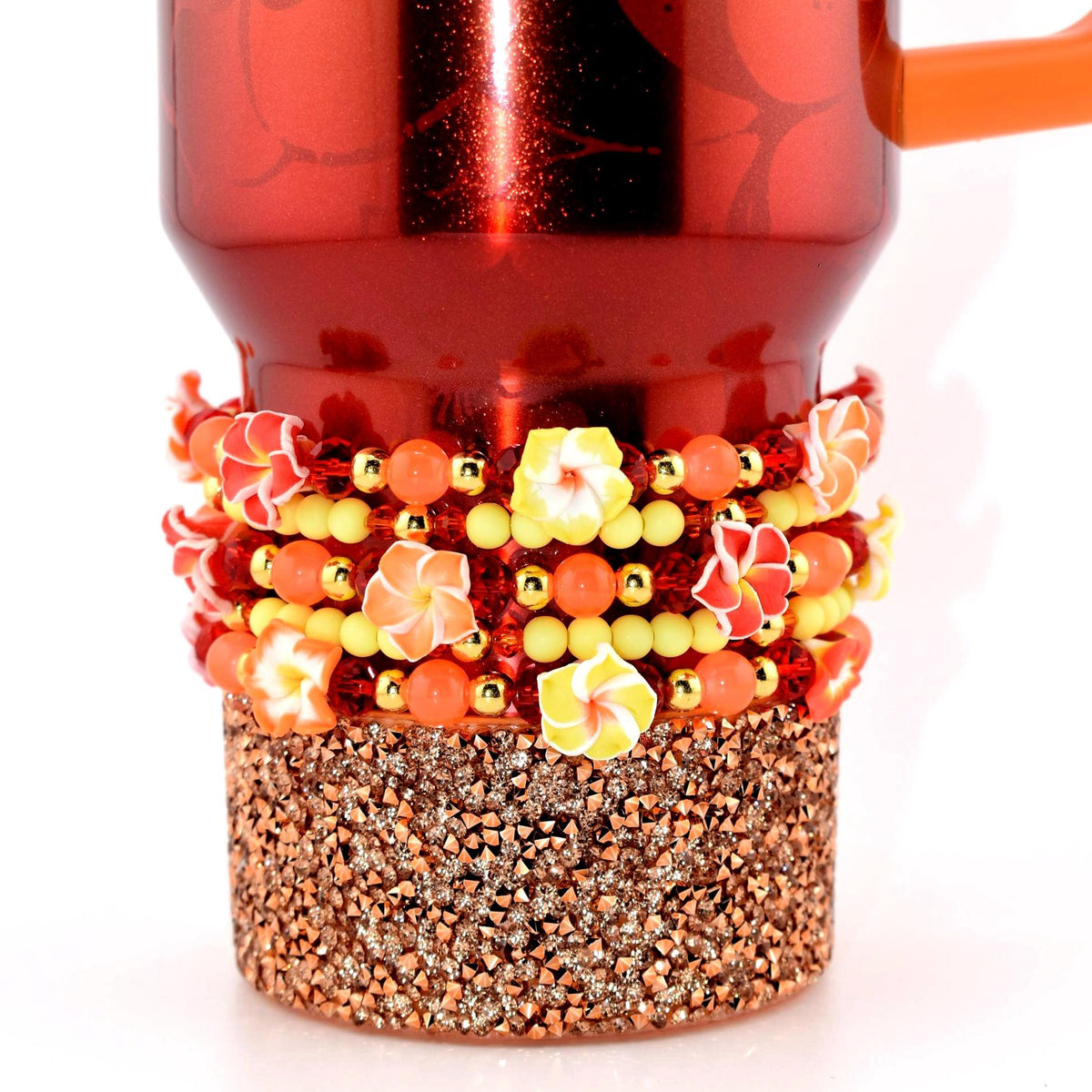 A red tumbler with an orange handle. The base is covered in sparkling copper-colored crystals. A beaded band with yellow, orange, and red floral accents encircles the mug.