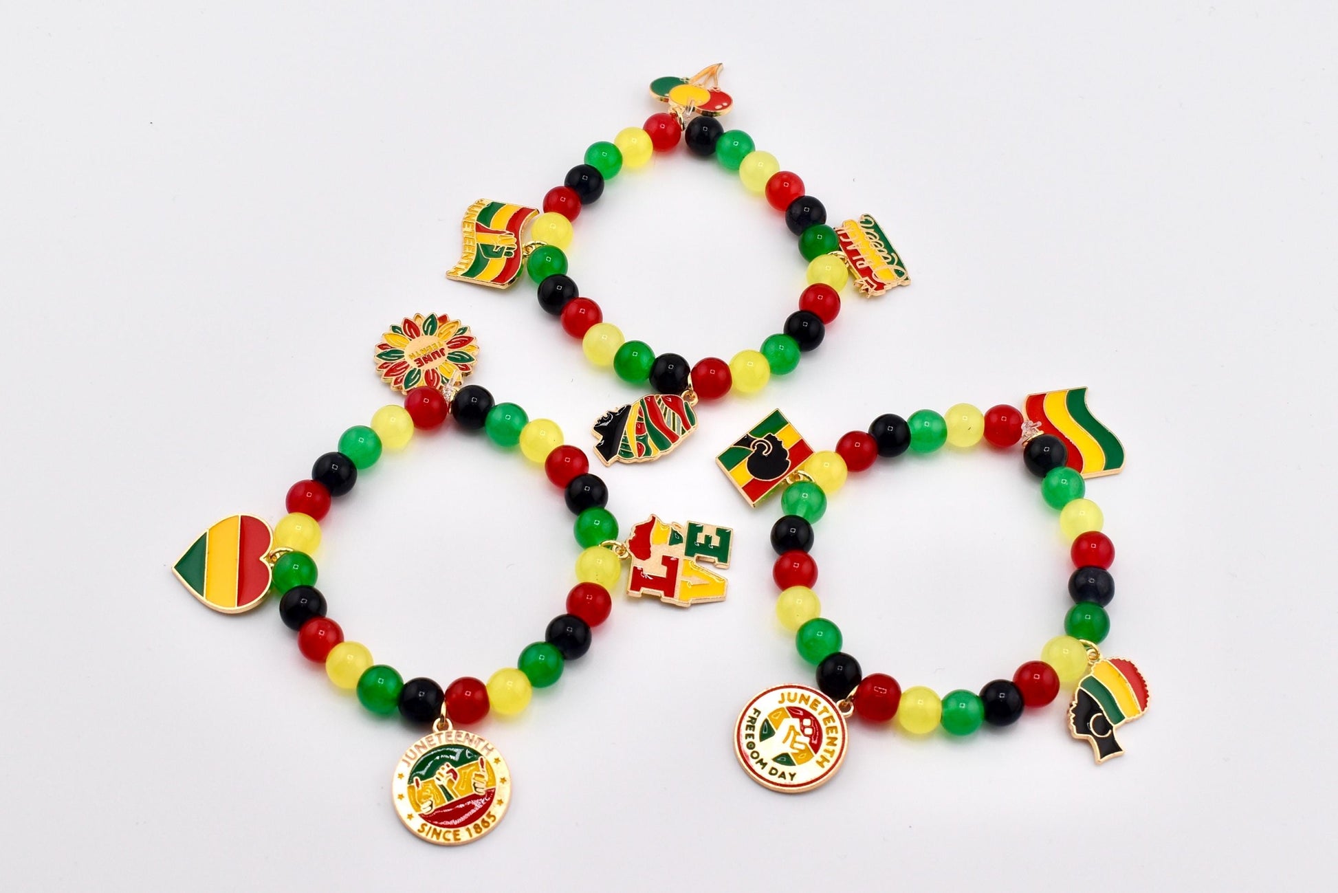 Three beaded bracelets with red, yellow, green, and black beads. Each bracelet features Juneteenth-themed charms, including flags, a heart, and a medallion with text. The bracelets are displayed on a white surface.