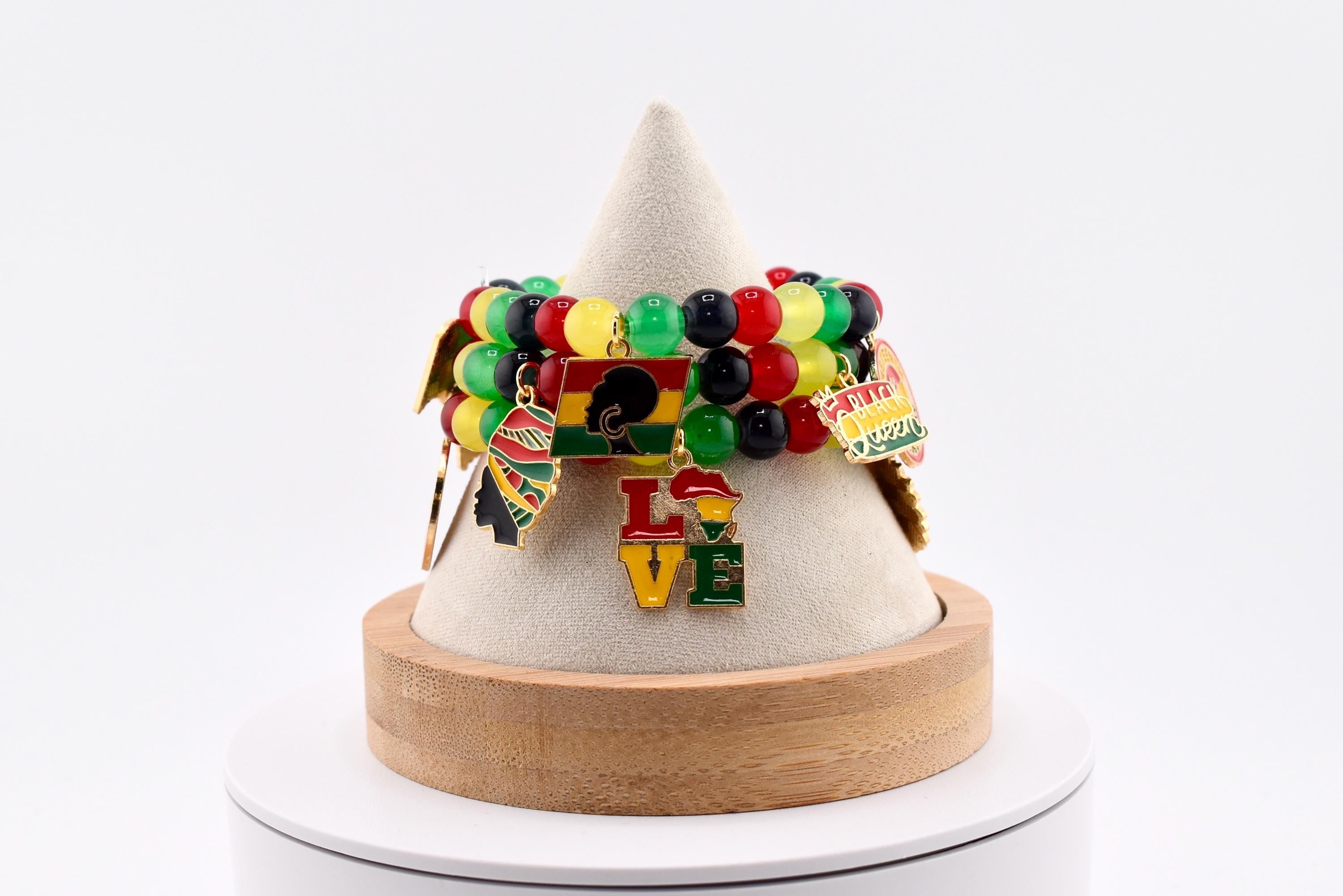 A beaded bracelet with red, yellow, green, and black beads. The bracelet has Juneteenth gold charms with the word "LOVE" and other cultural symbols. The bracelet is displayed on a white cone-shaped stand.