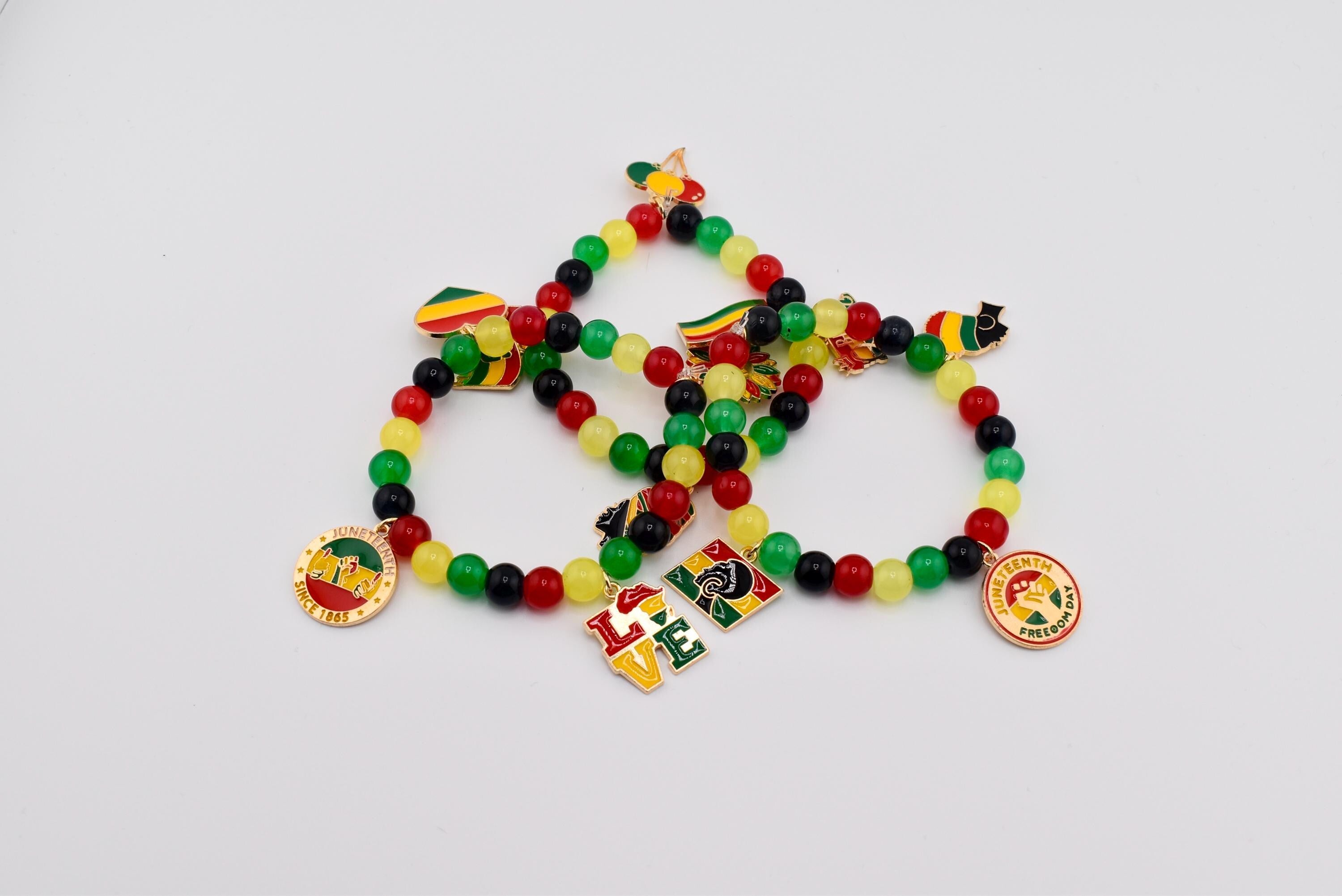 Three beaded bracelets with red, yellow, green, and black beads. Each bracelet has charms with flags, the word "LOVE", and a round charm with text. The bracelets are on a white background.