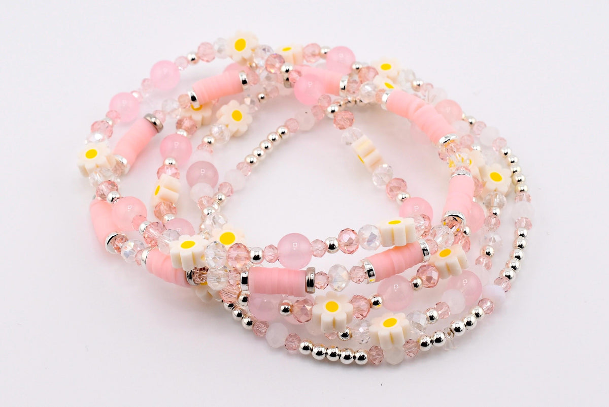 Several beaded bracelets in shades of pink, white, and silver. The bracelets feature a mix of beads, including clear crystal, pink round beads, and small white daisy-shaped beads with yellow centers. The bracelets are layered.