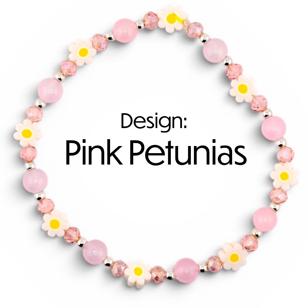 A pink beaded bracelet with the text "Design: Pink Petunias". The bracelet features alternating pink beads, clear faceted beads, and white daisy-shaped beads with yellow centers. The beads are strung on a silver-colored chain.
