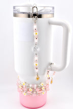 White tumbler with a pink base and a handle, adorned with a daisy-themed charm and bracelet. The charm features pink beads, white daisy accents, and a crystal pendant. The bracelet has pink and white beads.