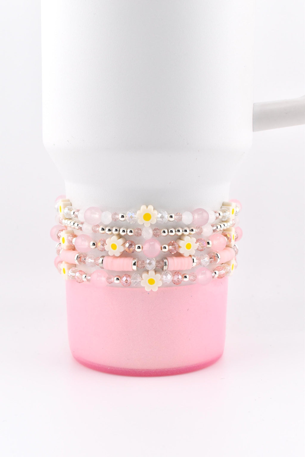 Stack of five beaded bracelets in shades of pink and white, adorned with small daisy accents. The bracelets are wrapped around a white mug with a pink base, creating a cohesive and stylish accessory.