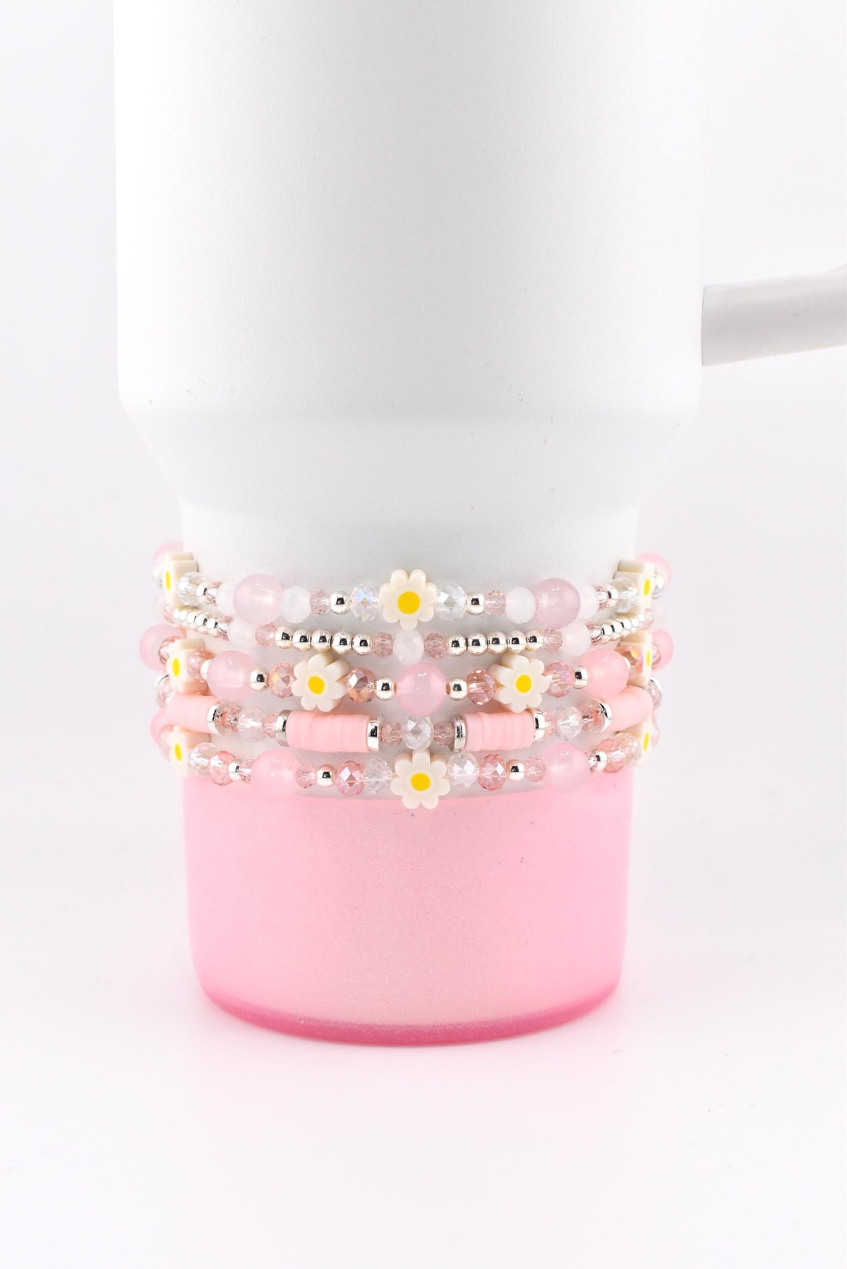 Stack of five beaded bracelets in shades of pink and white, adorned with small daisy accents. The bracelets are wrapped around a white mug with a pink base, creating a cohesive and stylish accessory.