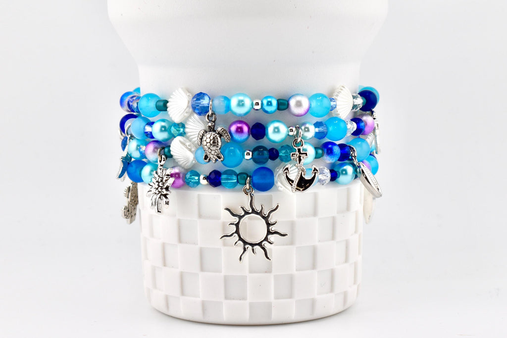 Several beaded bracelets in shades of blue, white, and purple are stacked on a white, textured surface. Each bracelet features various charms, including a sun, turtle, and anchor. The bracelets are adorned with beads and seashell accents.