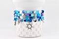 Several beaded bracelets in shades of blue, white, and purple are stacked on a white, textured surface. Each bracelet features various charms, including a sun, turtle, and anchor. The bracelets are adorned with beads and seashell accents.