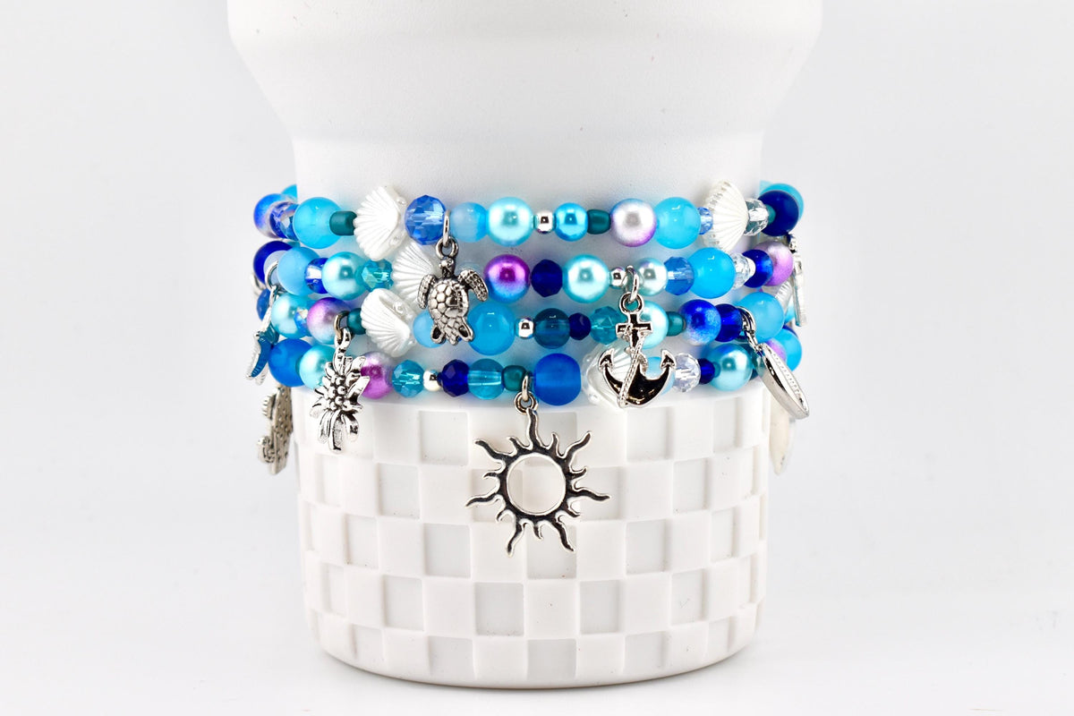 Several beaded bracelets in shades of blue, white, and purple are stacked on a white, textured surface. Each bracelet features various charms, including a sun, turtle, and anchor. The bracelets are adorned with beads and seashell accents.