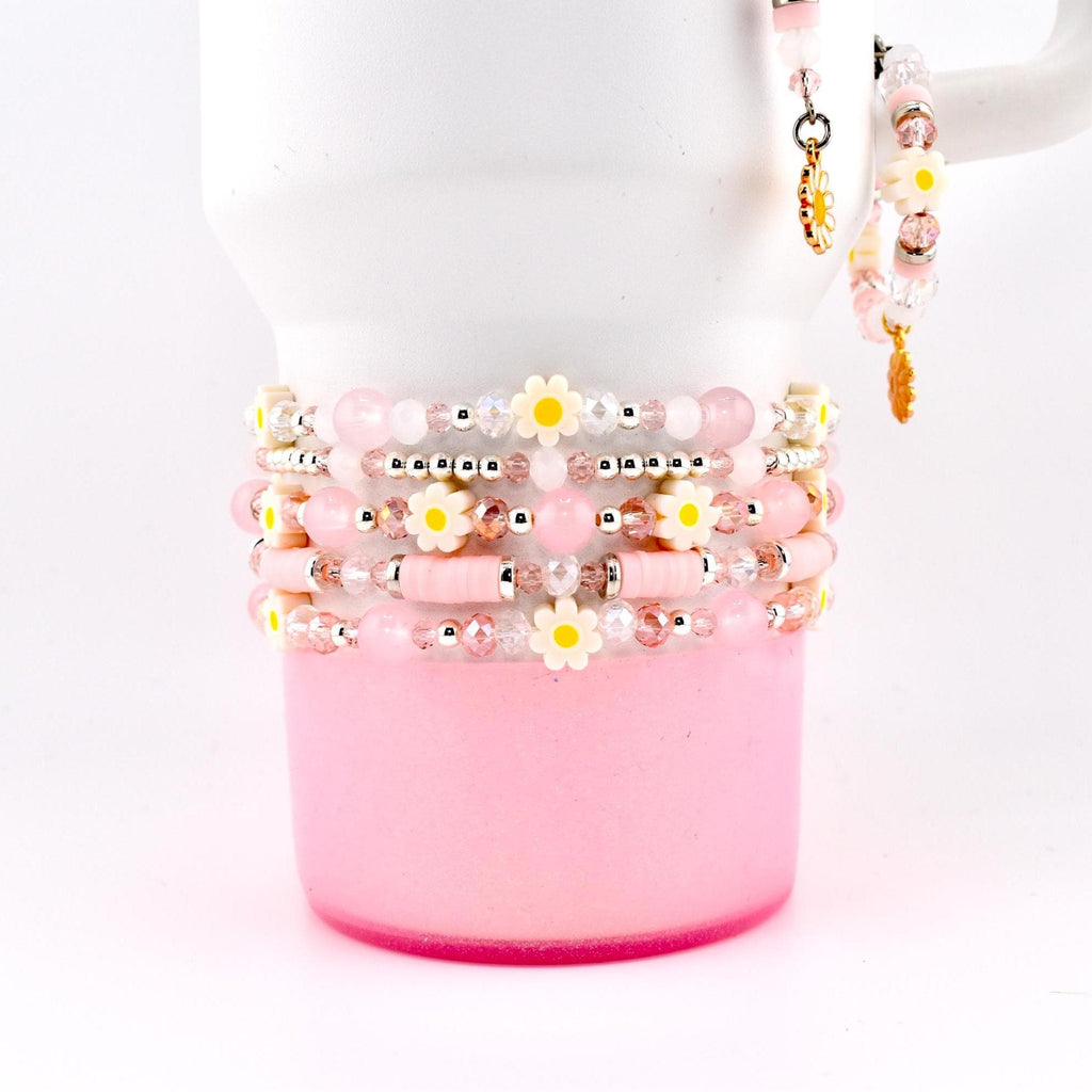 Stack of pink and white beaded bracelets with daisy accents. The bracelets are wrapped around a white mug with a pink base. A matching bracelet and charm hang from the mug's handle. The bracelets feature various bead sizes and shapes.