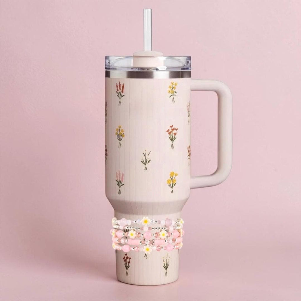A light pink insulated tumbler with a handle, lid, and straw. The tumbler is decorated with small floral bouquets. A stack of beaded bracelets in shades of pink and white is wrapped around the base of the tumbler.