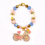 A beaded tumbler handle bracelet with a gold-colored bicycle charm. The bracelet features pink, blue, and clear beads, with gold accents and a lobster clasp. The bicycle charm has a pink seat and a basket of flowers.