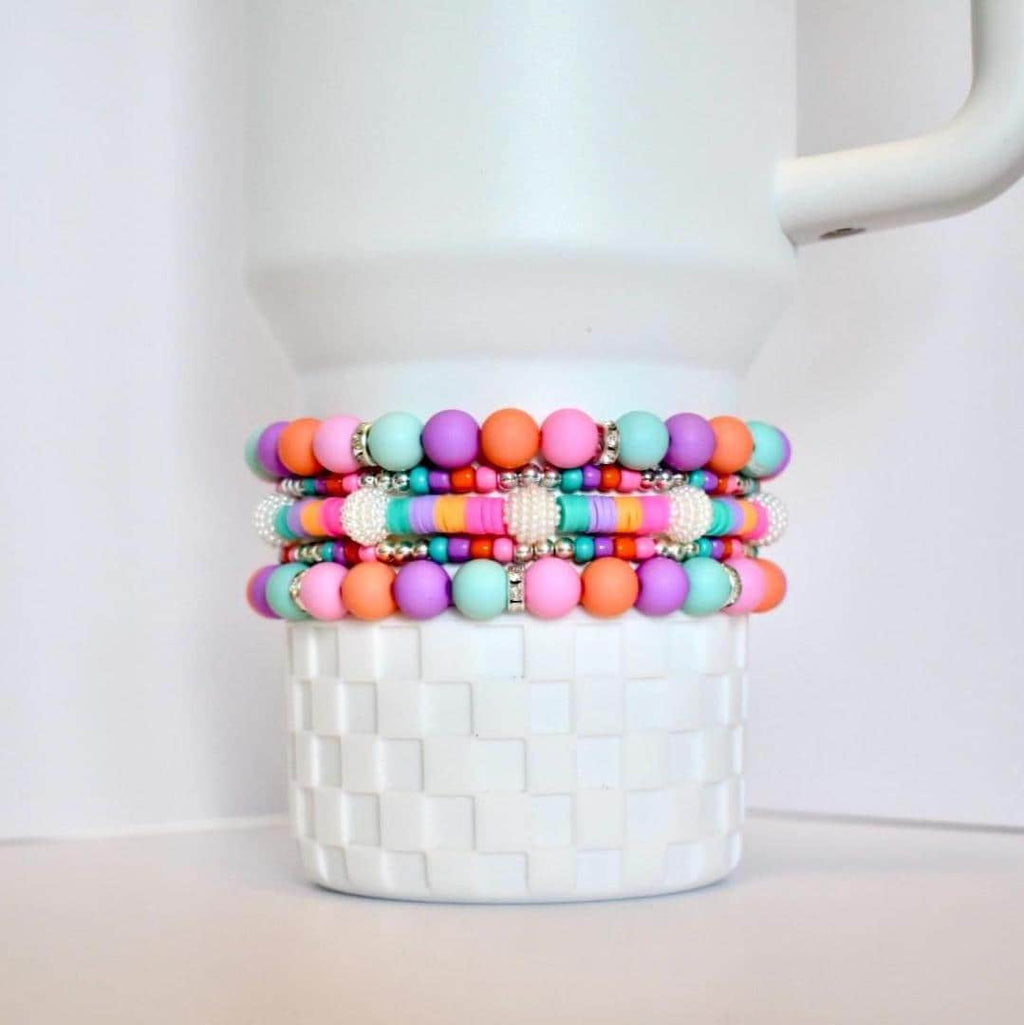 Four colorful beaded bracelets in shades of pink, orange, purple, and turquoise. The bracelets are stacked on top of a white, textured container. Each bracelet has silver accents.