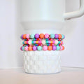 Four colorful beaded bracelets in shades of pink, orange, purple, and turquoise. The bracelets are stacked on top of a white, textured container. Each bracelet has silver accents.