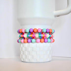 Four colorful beaded bracelets in shades of pink, orange, purple, and turquoise. The bracelets are stacked on top of a white, textured container. Each bracelet has silver accents.
