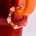 A beaded tumbler handle bracelet with orange, red, and gold accents. The bracelet features small white and red flower-shaped beads, interspersed with orange and red crystal beads and gold-colored beads. The bracelet is attached to an orange Stanley handle.
