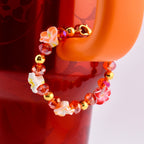 A beaded tumbler handle bracelet with orange, red, and gold accents. The bracelet features small white and red flower-shaped beads, interspersed with orange and red crystal beads and gold-colored beads. The bracelet is attached to an orange Stanley handle.