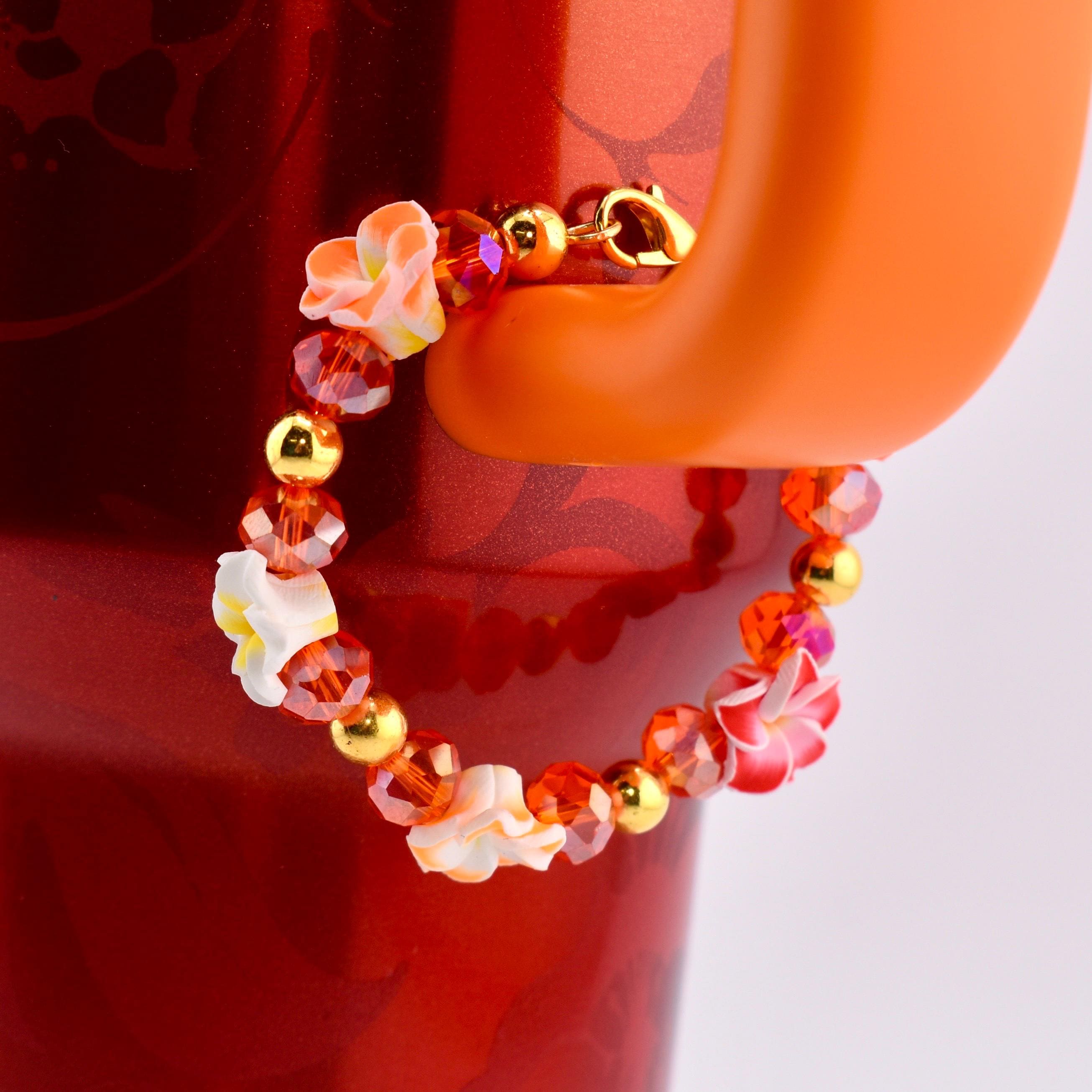 A beaded tumbler handle bracelet with orange, red, and gold accents. The bracelet features small white and red flower-shaped beads, interspersed with orange and red crystal beads and gold-colored beads. The bracelet is attached to an orange Stanley handle.