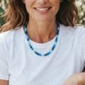 A beaded necklace with various shades of blue and green beads. The necklace is composed of round and cylindrical beads, creating a colorful and textured accessory. The necklace is worn against a white t-shirt.