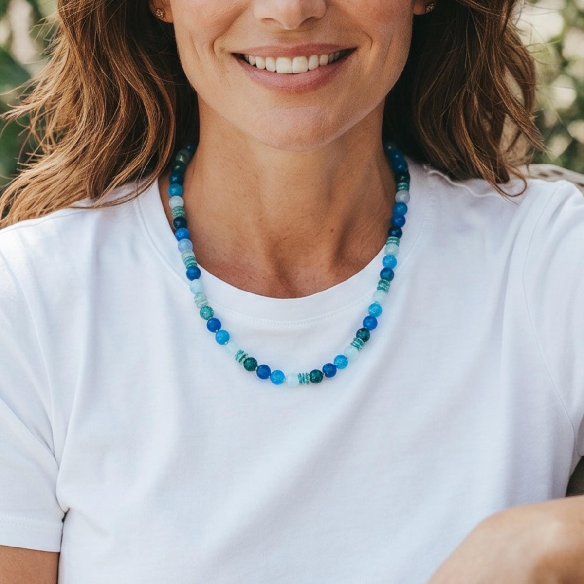 A beaded necklace with various shades of blue and green beads. The necklace is composed of round and cylindrical beads, creating a colorful and textured accessory. The necklace is worn against a white t-shirt.