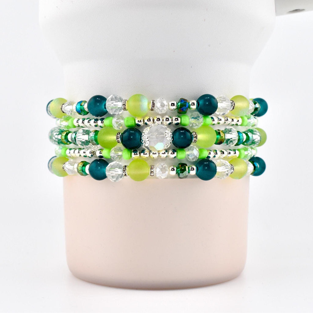 Stack of five beaded bracelets in shades of green, teal, and clear. The bracelets feature a variety of bead sizes and shapes, including round, faceted, and crystal beads. The bracelets are displayed on a white cylindrical object.