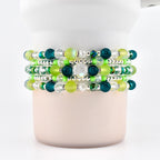 Stack of five beaded bracelets in shades of green, teal, and clear. The bracelets feature a variety of bead sizes and shapes, including round, faceted, and crystal beads. The bracelets are displayed on a white cylindrical object.