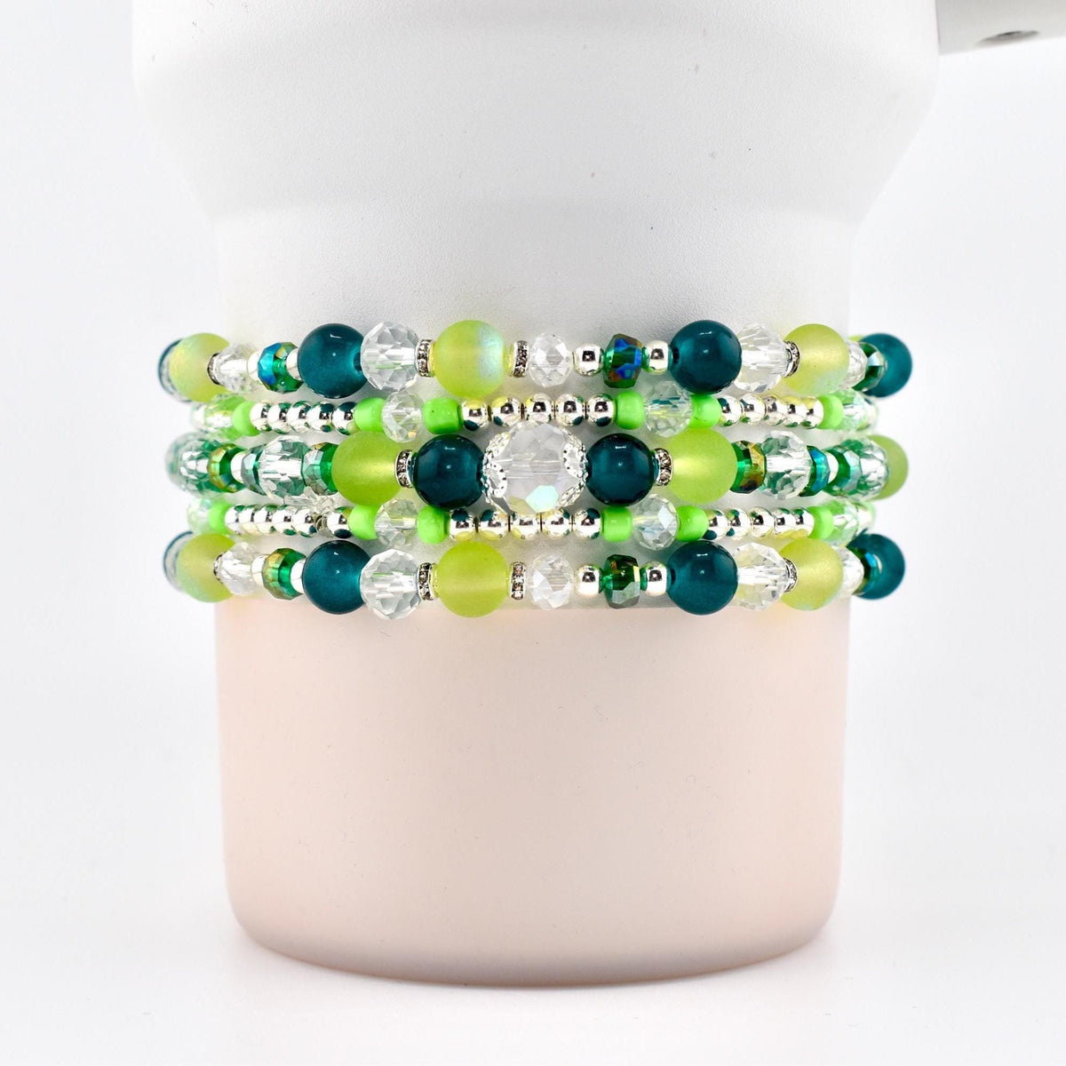 Stack of five beaded bracelets in shades of green, teal, and clear. The bracelets feature a variety of bead sizes and shapes, including round, faceted, and crystal beads. The bracelets are displayed on a white cylindrical object.